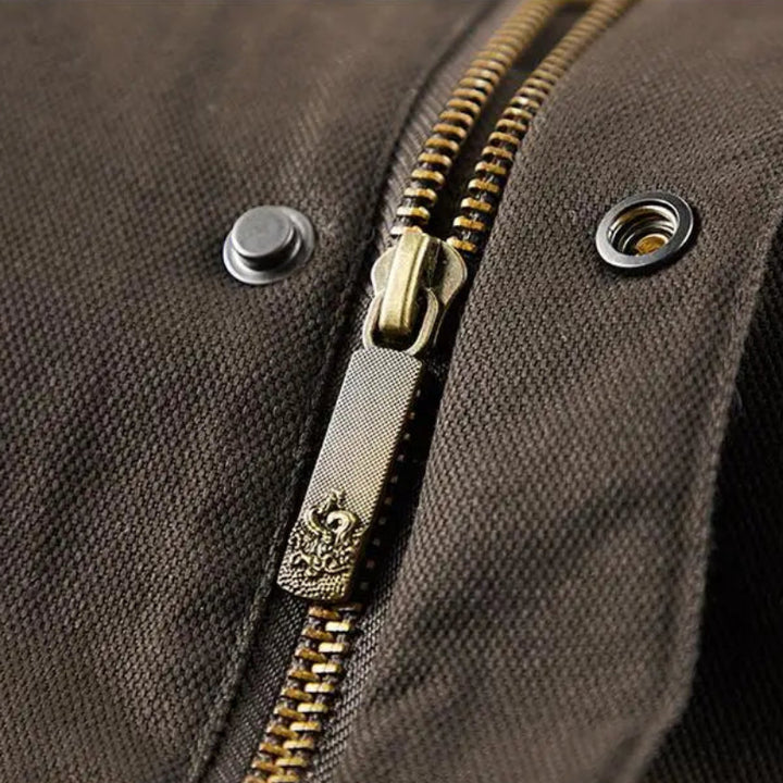 Close-up of a dark brown jacket fabric with an antique brass zipper pull featuring an ornate crest design.