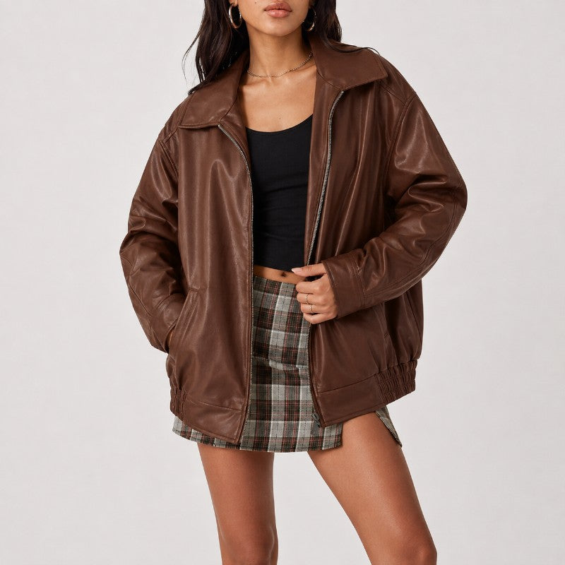 A woman wearing an oversized brown faux-leather jacket over a black tank top and a brown plaid mini skirt, photographed against a light gray studio background.