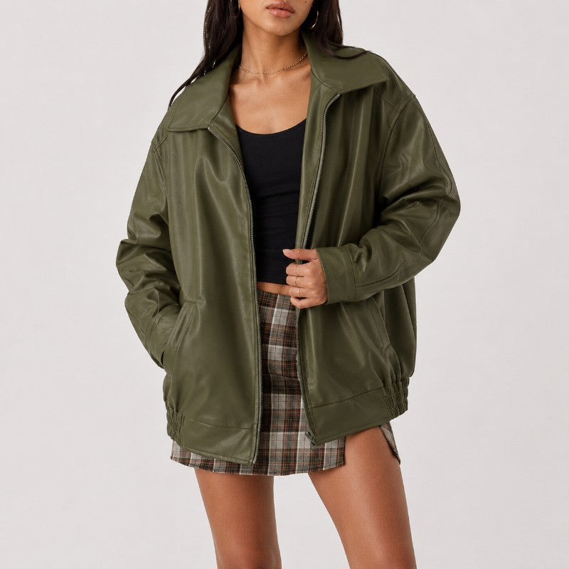 A woman wearing an oversized olive green faux-leather jacket over a black tank top and a brown plaid mini skirt, photographed against a light gray studio background.