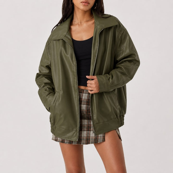 A woman wearing an oversized olive green faux-leather jacket over a black tank top and a brown plaid mini skirt, photographed against a light gray studio background.