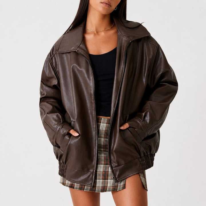 A woman wearing an oversized dark brown faux-leather jacket with a high collar, front zipper, and side pockets, styled over a black top and a plaid mini skirt against a plain light gray background.