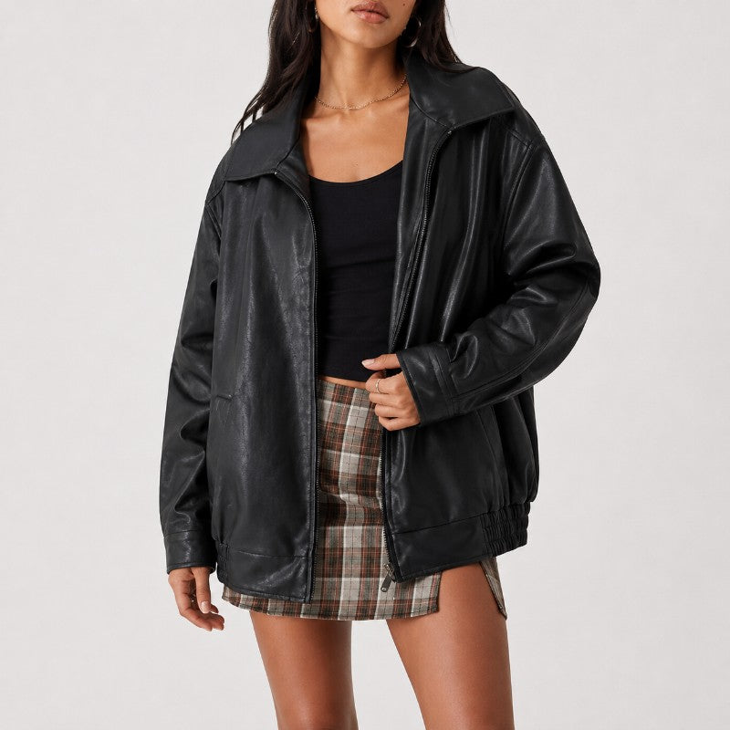 A woman wearing an oversized black faux-leather jacket over a black tank top and a brown plaid mini skirt, photographed against a light gray studio background.
