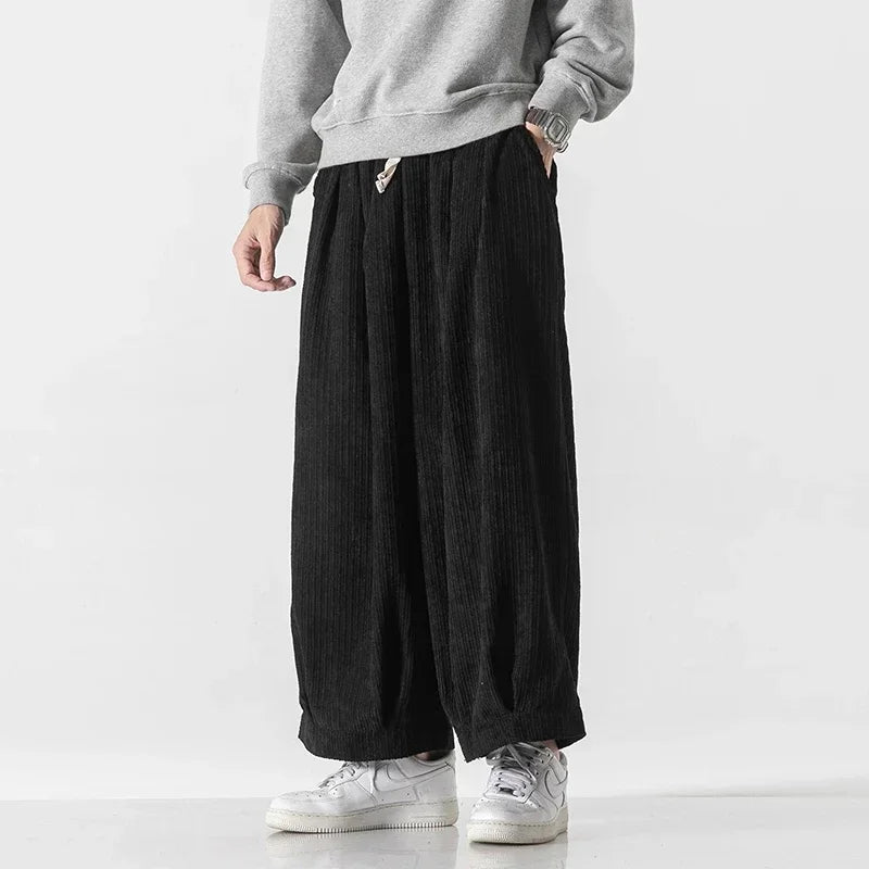 Model wearing oversized black corduroy pants with deep pleats and elastic waistband for comfortable fit.