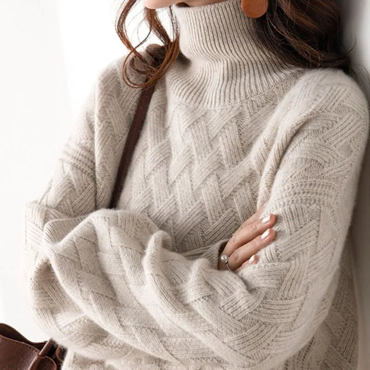 A close-up of a person wearing a chunky knit turtleneck sweater in a warm off-white or cream tone. The sweater has a textured cable and basketweave pattern, and the person is shown from the neck down with arms crossed, wearing a brown shoulder bag and ring.