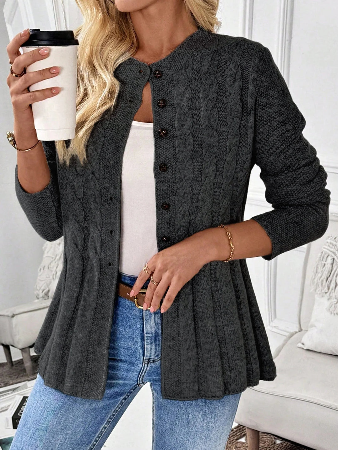 Woman wearing a dark gray cable knit cardigan with brown buttons, a peplum hem, light wash jeans, and holding a coffee cup.