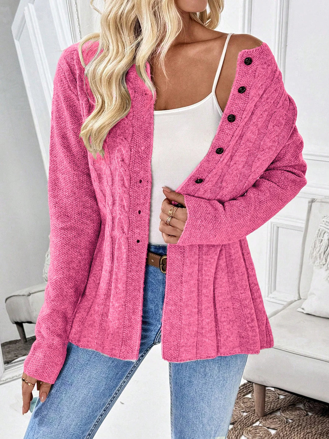 Woman wearing an open bright pink cable knit cardigan with black buttons, over a white tank top and blue jeans.