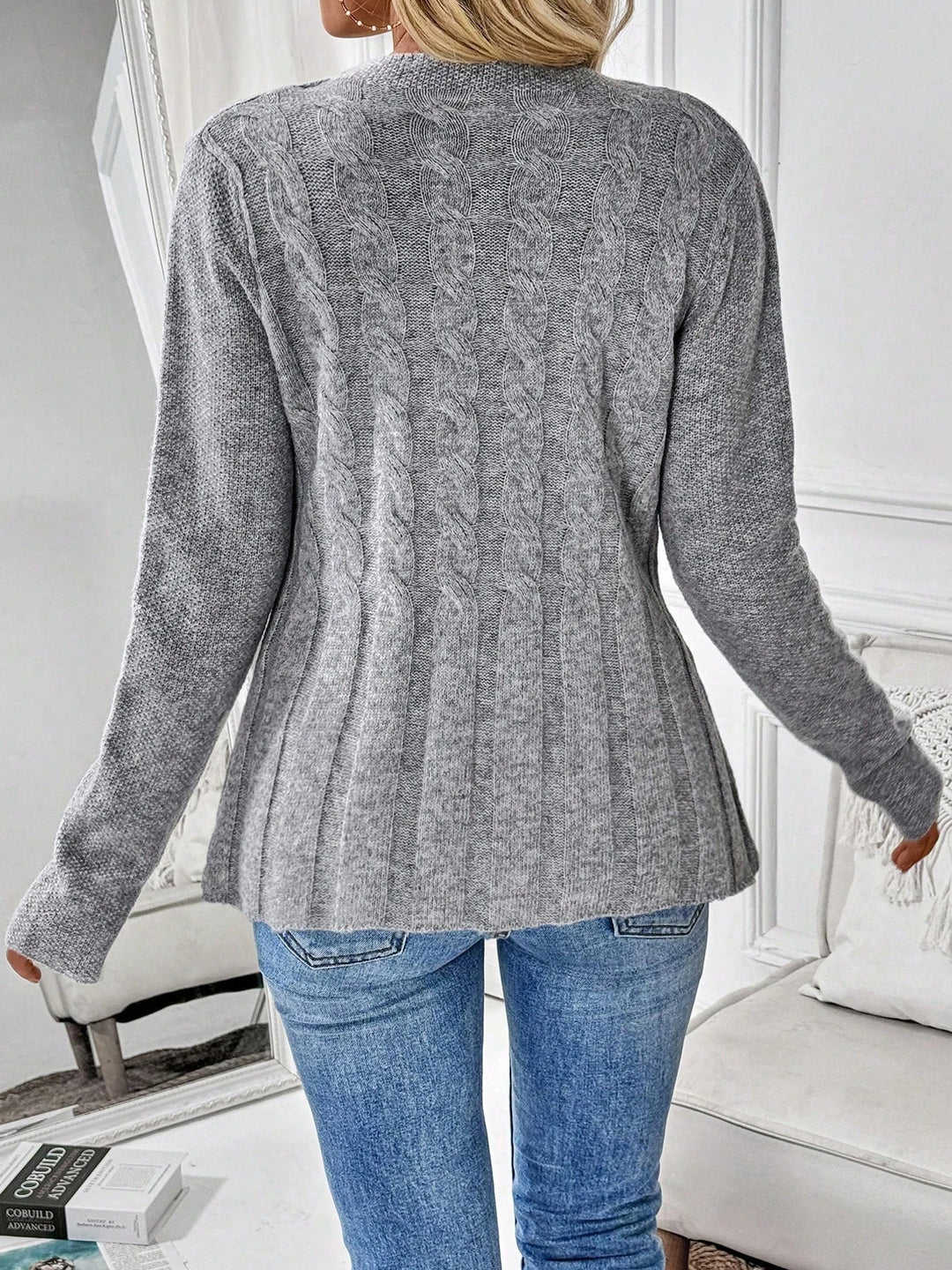 Woman wearing a gray cable knit cardigan with ribbed sleeves and a pleated hem, paired with light blue jeans.