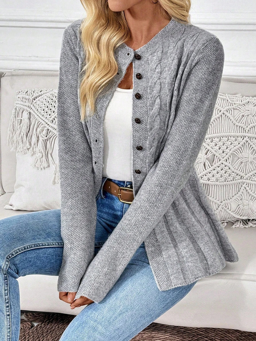 Woman wearing a light gray knit cardigan with brown buttons, a white tank top, and blue jeans, sitting on a couch.