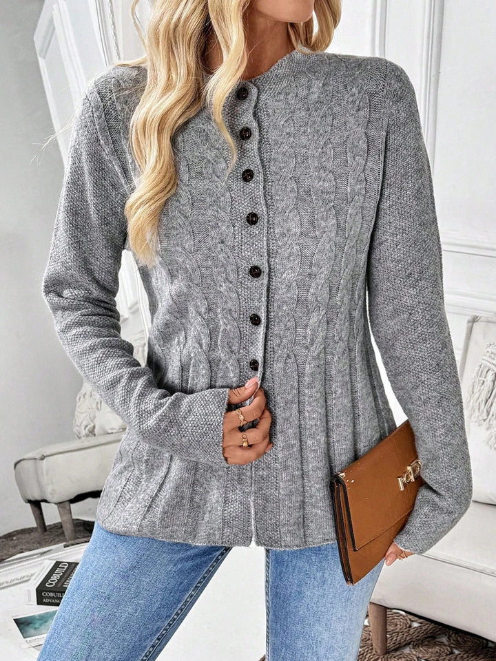 Woman wearing a light gray cable-knit cardigan with dark buttons, blue jeans, and holding a brown clutch, indoors.