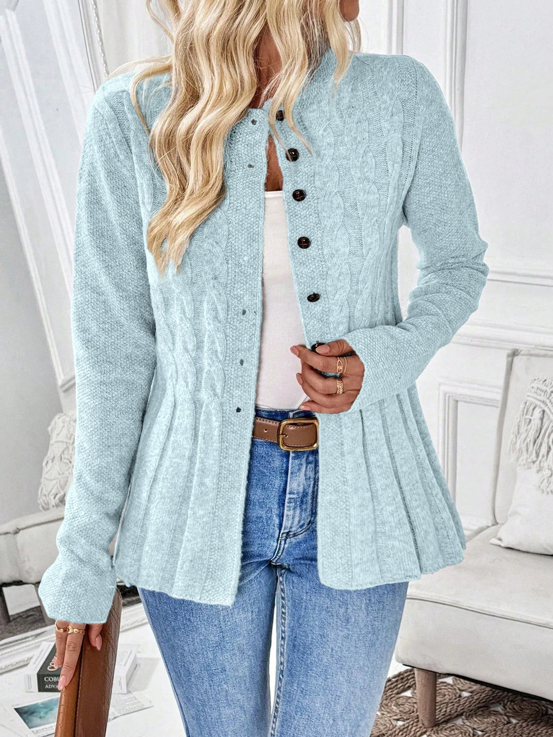 Woman wearing a light blue cable knit cardigan with dark brown buttons, a pleated hem, jeans, and a brown belt.