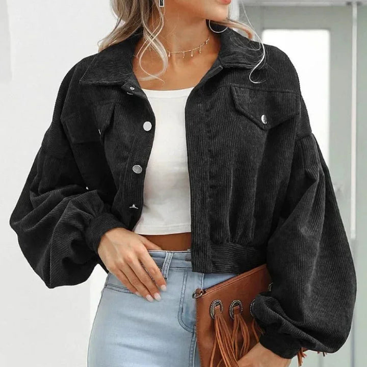 Woman wearing a black corduroy cropped jacket with puffed sleeves, silver buttons, and chest pockets over a white top.