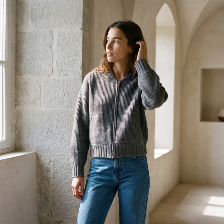 Woman wearing a gray ribbed knit zip-up cardigan and blue jeans, looking out a window in a stone room.