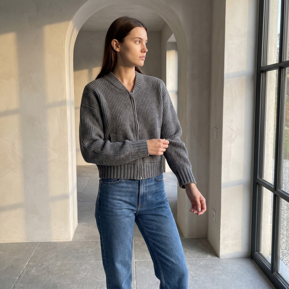 Woman wearing a dark gray ribbed knit zip-up cardigan with a collarless neckline and medium-wash blue jeans, looking out a large window.
