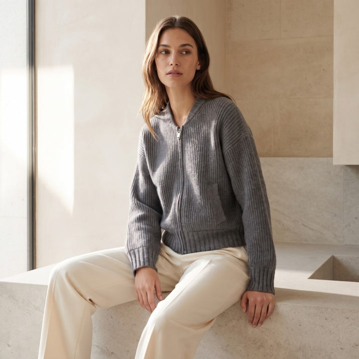 Woman wearing a gray ribbed knit zip-up cardigan with a silver zipper and light beige pants, sitting indoors.