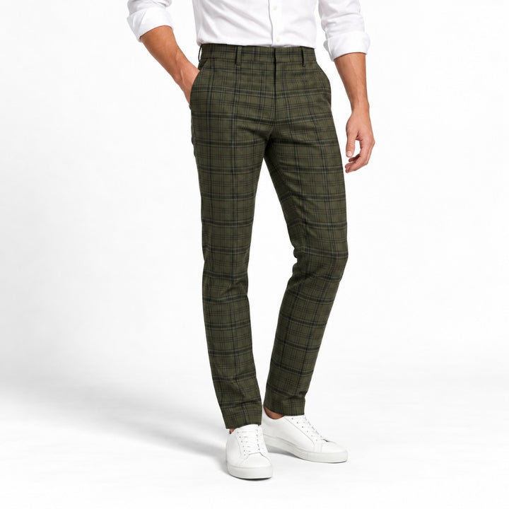 Man wearing olive green plaid slacks with a slim fit, a white dress shirt, and white sneakers, posing with one hand in a pocket.