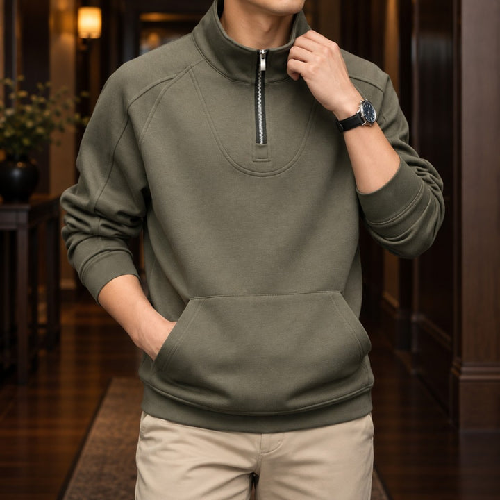 Man wearing an olive green quarter-zip pullover with raglan sleeves and a front kangaroo pocket, standing indoors.