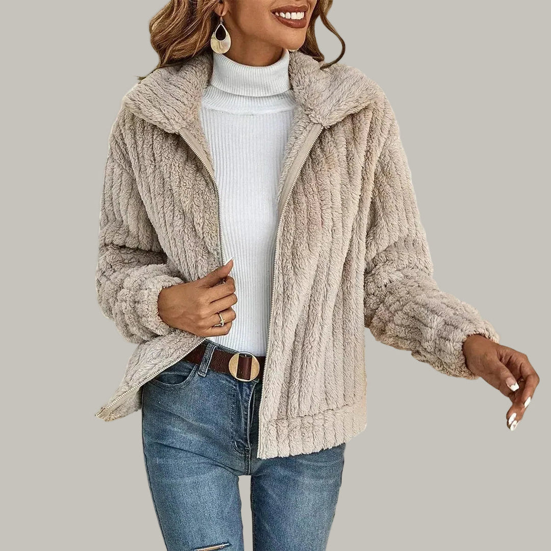 Woman wearing an open, textured beige fleece jacket over a white ribbed turtleneck and blue jeans with a brown belt.