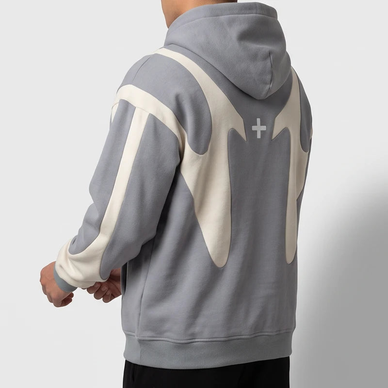 Man wearing an oversized grey hoodie featuring cream curved back paneling and a small white cross against a white backdrop.