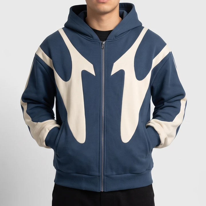 Man wearing a slate blue zip-up hoodie featuring a bold cream abstract design, posed with hands in his pockets.