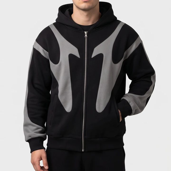 Man wearing a black zip-up hoodie with contrasting grey flame-shaped panels across the chest and sleeves against a white backdrop.