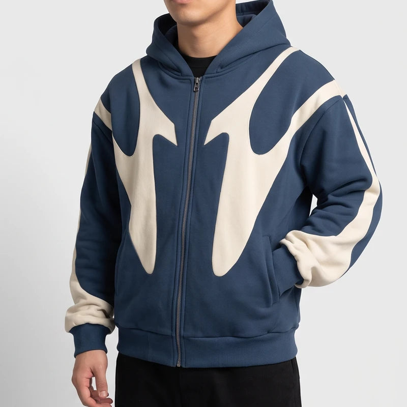 Man wearing a slate blue zip-up hoodie with cream abstract panels, posing with one hand in his pocket.