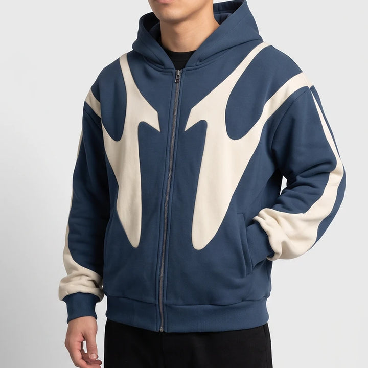 Man wearing a slate blue zip-up hoodie with cream abstract panels, posing with one hand in his pocket.