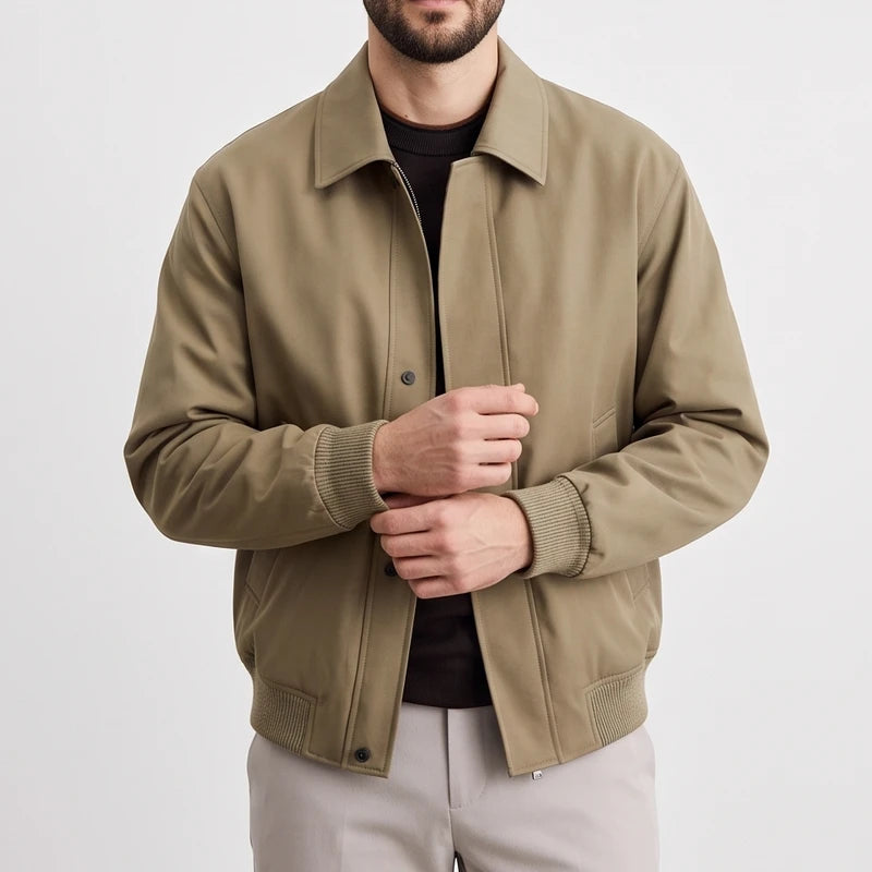 Man wearing an open light brown bomber jacket with ribbed cuffs and hem, over a dark shirt and light pants.