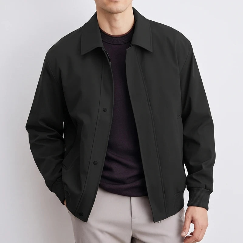 Man wearing a black blouson jacket with a collar, ribbed cuffs, and button placket over a dark sweater and light grey pants.