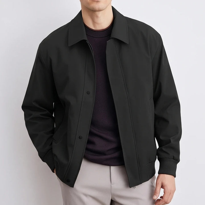 Man wearing a black blouson jacket with a collar, ribbed cuffs, and button placket over a dark sweater and light grey pants.