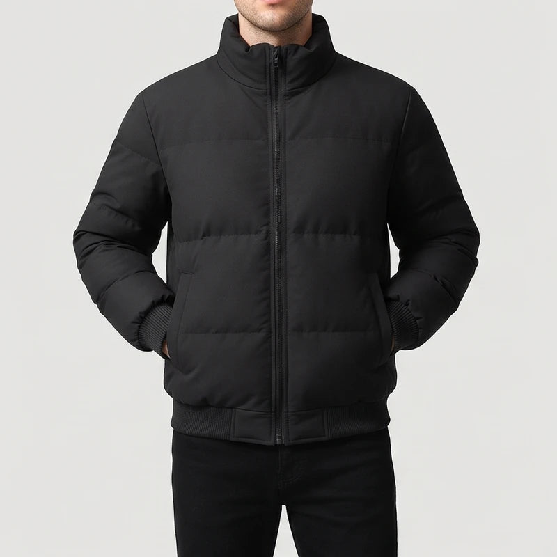 Man wearing a black puffer jacket with a high collar and full zipper, hands in pockets, against a light background.