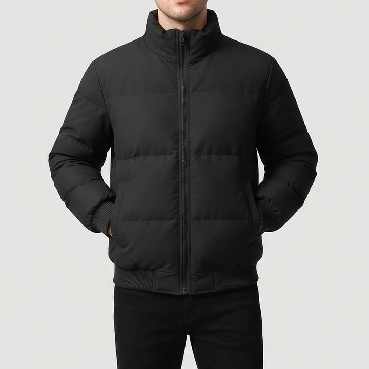 Man wearing a black puffer jacket with a high collar and full zipper, hands in pockets, against a light background.