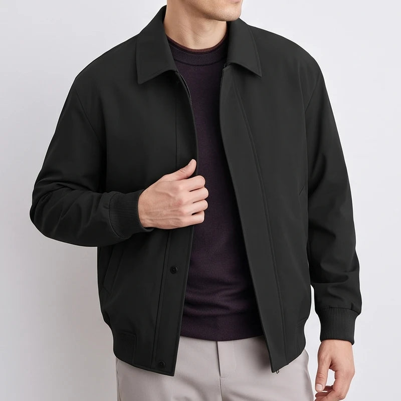Man wearing a black collared bomber jacket with ribbed cuffs and hem, partially open over a dark sweater and light pants.