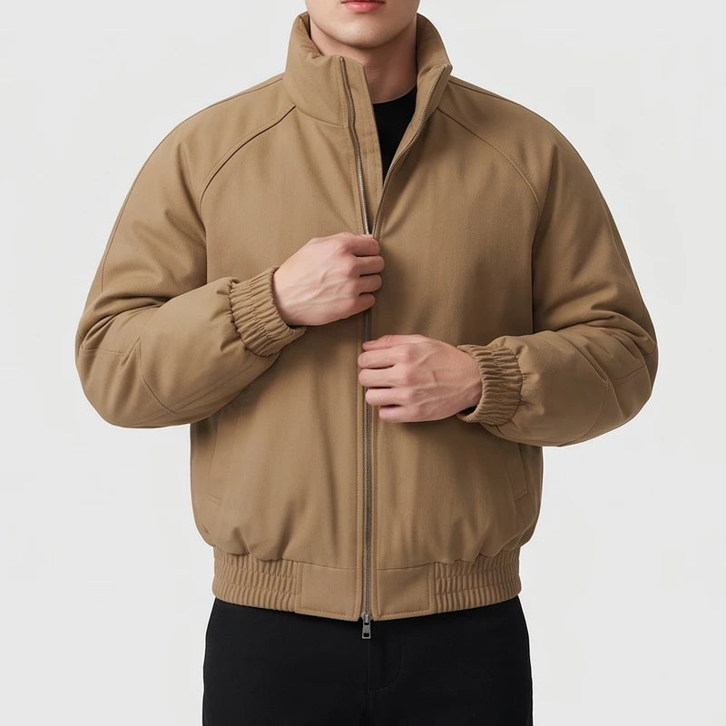 Man wearing a tan bomber jacket with a high collar, ribbed cuffs, and hem, zipping it up against a white background.