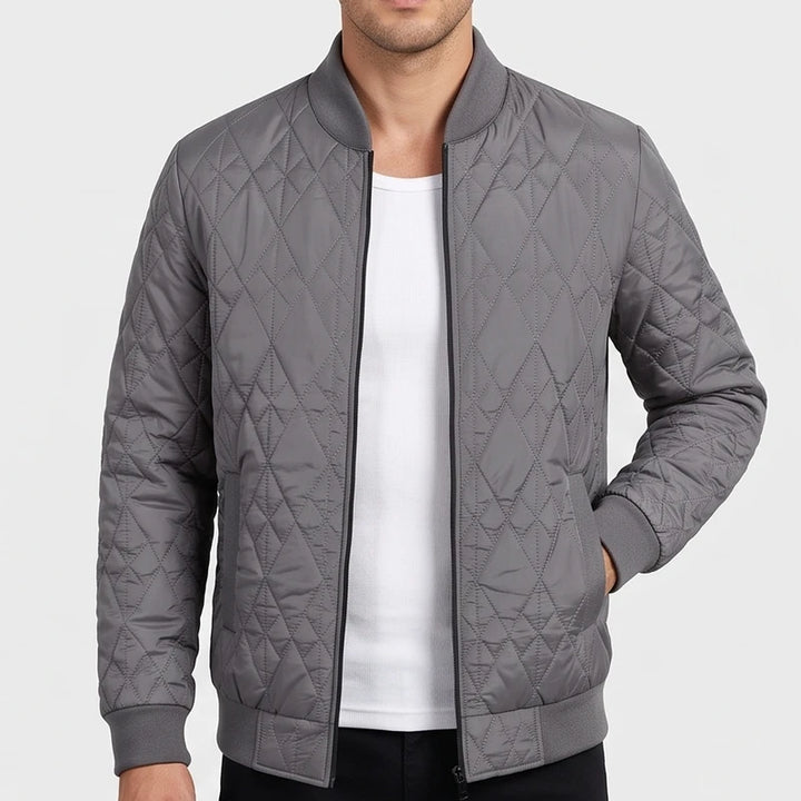 Man wearing a grey quilted bomber jacket with a diamond pattern, black zipper, and ribbed trim, with his hand in a pocket.