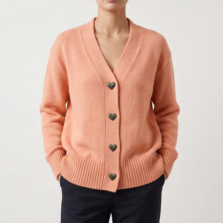 Woman wearing a peach knit cardigan with a V-neck, textured heart-shaped buttons, and ribbed cuffs and hem.