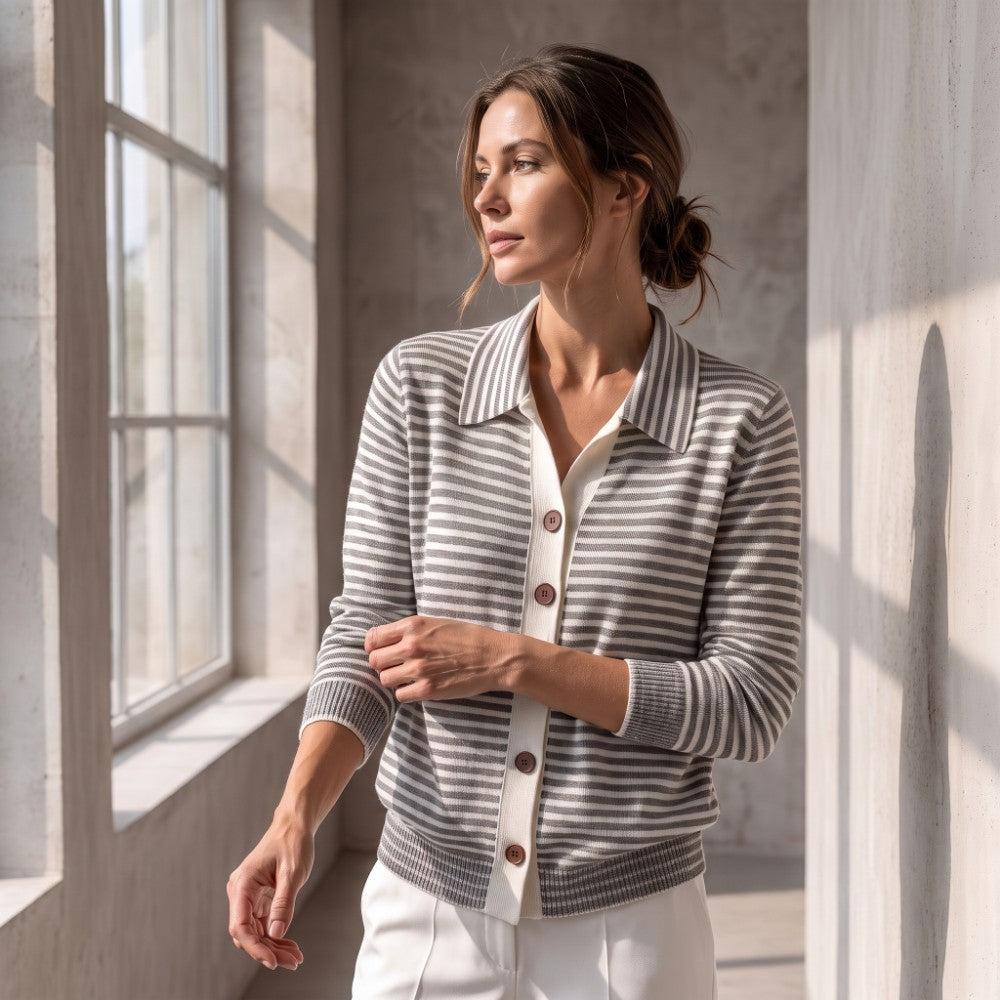 Woman wearing a grey and white striped knit cardigan with a ribbed collar and five brown buttons, standing by a window.