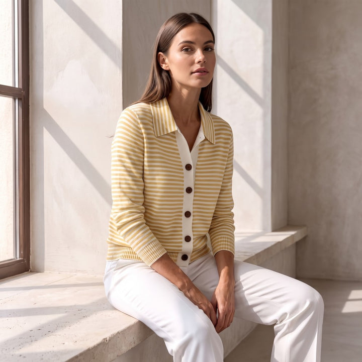 Woman wearing a yellow and white striped knit cardigan with a collar, brown buttons, and white trousers, sitting by a window.