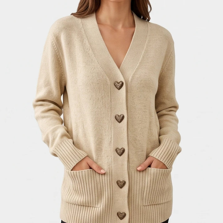 Woman wearing a beige knit cardigan with a V-neck, heart-shaped buttons, and ribbed pockets, hands in pockets.