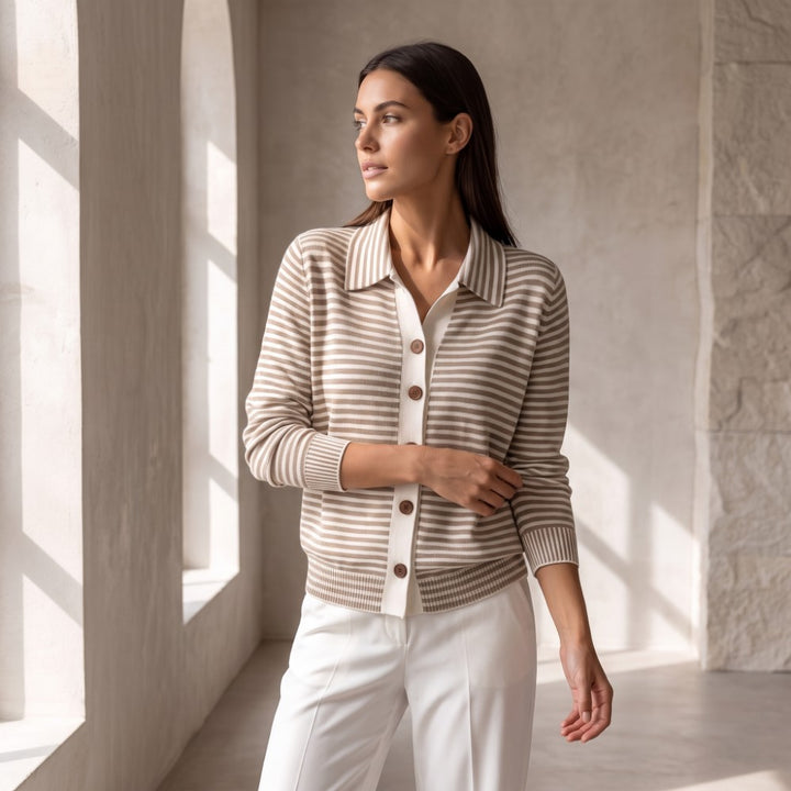 Woman looking left, wearing a collared, short beige and white striped knit top with brown buttons and white trousers.