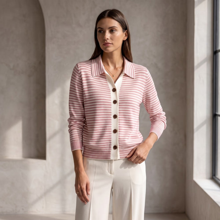 Woman standing and looking left, wearing a pink and white striped knit cardigan with a collar and brown buttons over white pants.