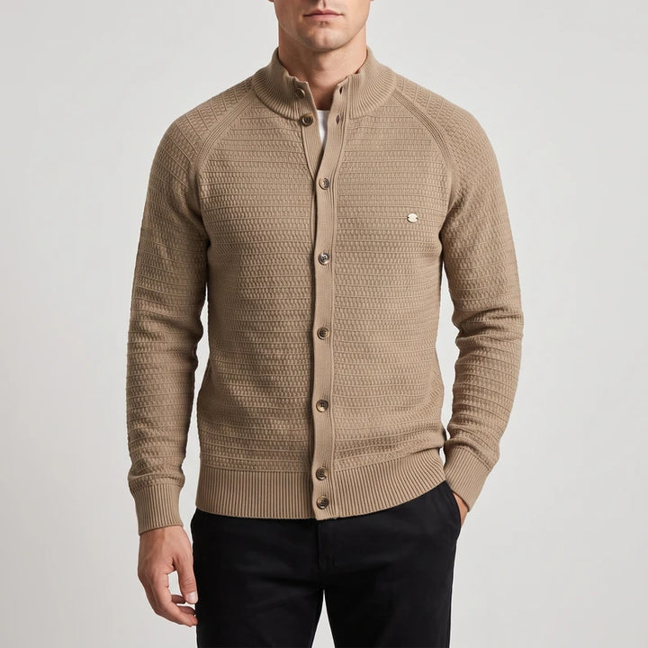 Man wearing a tan knit cardigan with a textured pattern, stand-up collar, and brown buttons, posing with one hand in his pocket.