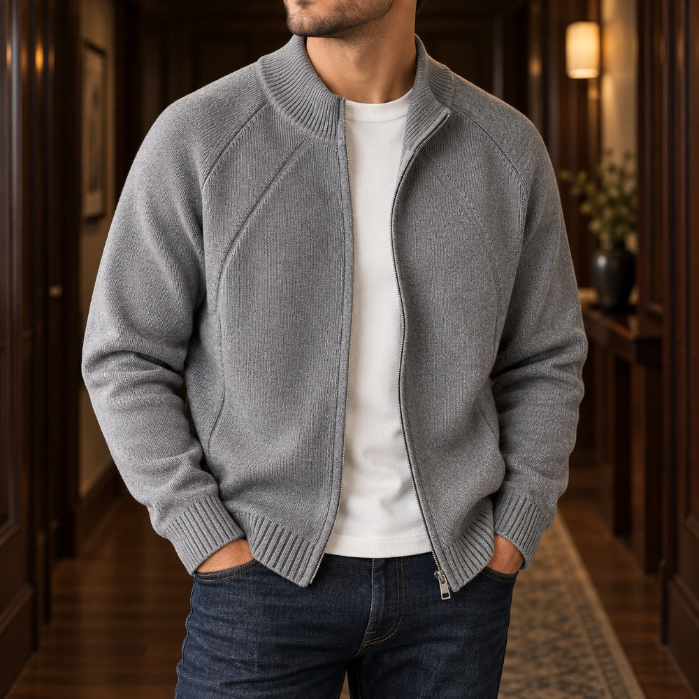 Man wearing a partially unzipped gray knit cardigan sweater over a white t-shirt, with hands in jeans pockets, standing in a hallway.