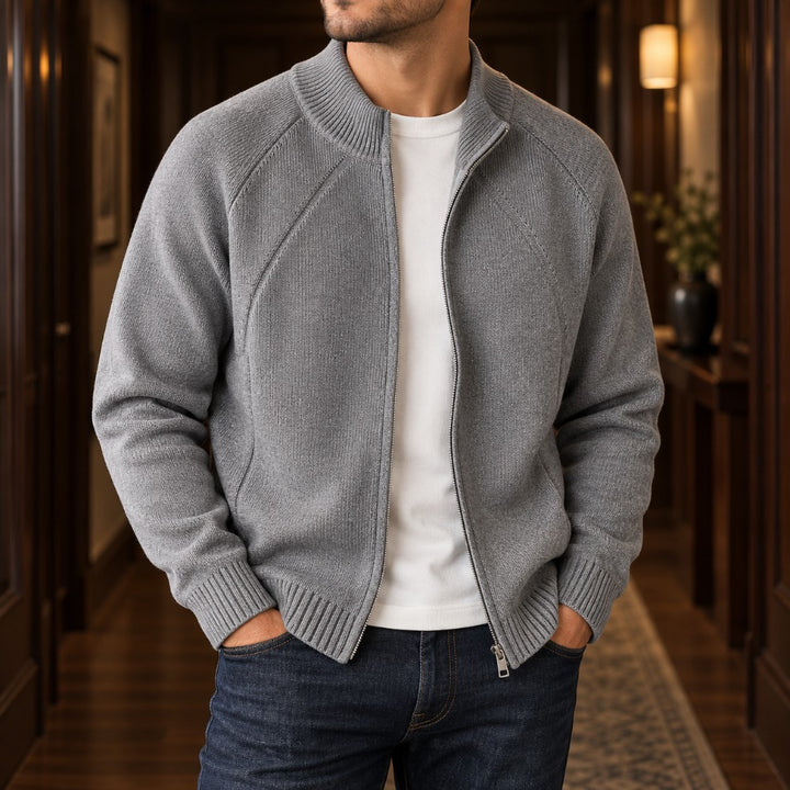 Man wearing a partially unzipped gray knit cardigan sweater over a white t-shirt, with hands in jeans pockets, standing in a hallway.