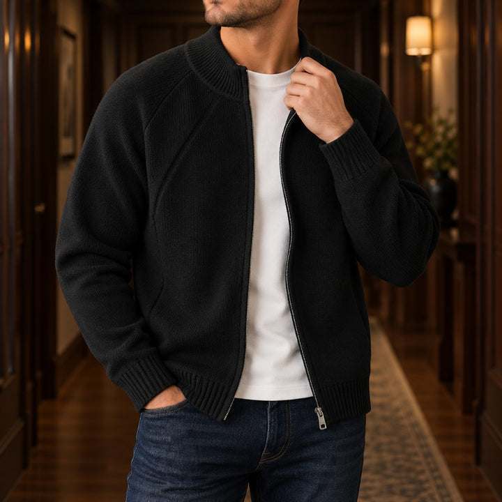 Man wearing an unzipped black knit sweater over a white t-shirt and dark blue jeans, standing in a hallway.