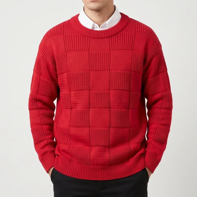Man wearing a vibrant red checkerboard knit sweater over a white collared shirt, hands in black pants pockets.