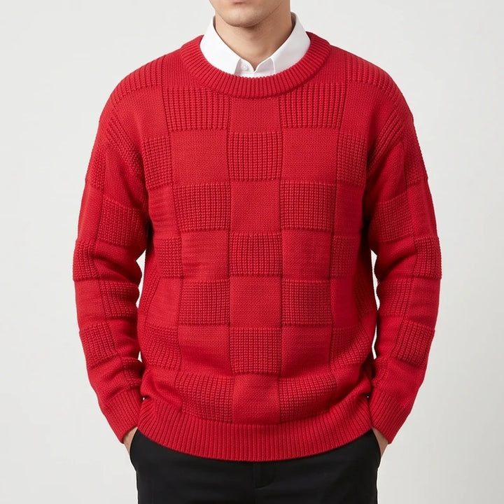 Man wearing a vibrant red checkerboard knit sweater over a white collared shirt, hands in black pants pockets.