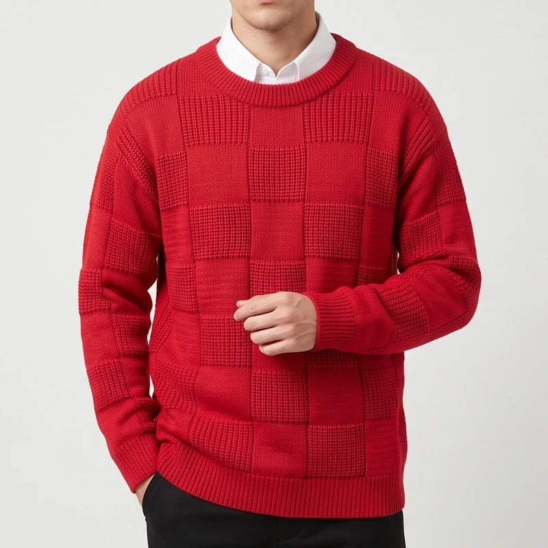 Man wearing a vibrant red checkerboard knit crew neck sweater over a white collared shirt, with his hand resting.