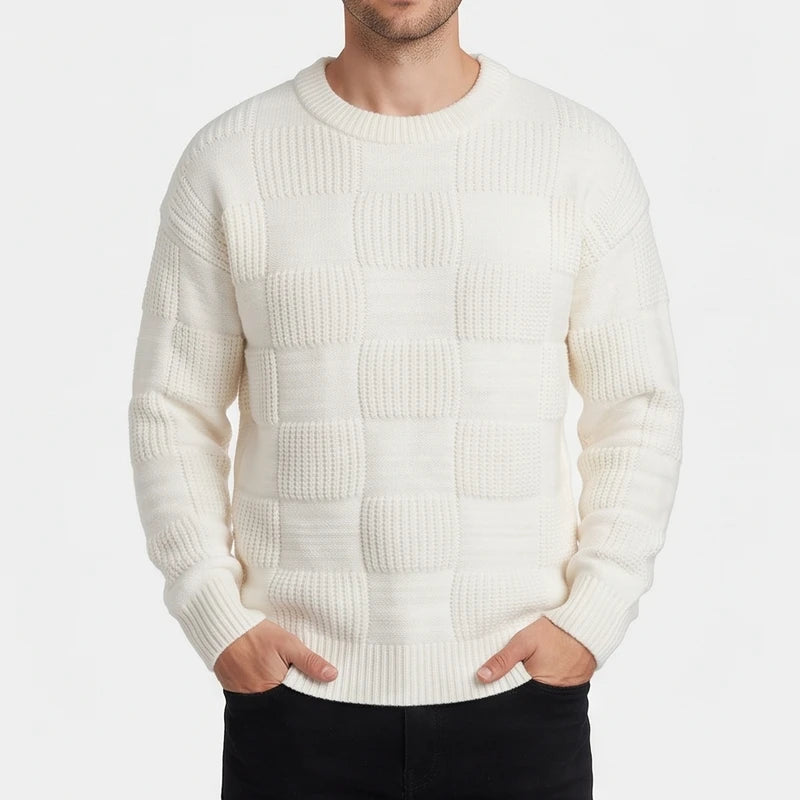 Man wearing an off-white knit sweater with a checkerboard texture, posing with hands in black jeans pockets.