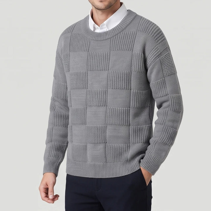 Man wearing a light gray knit crew neck sweater with a checkerboard textured pattern over a white collared shirt, hand in pocket.