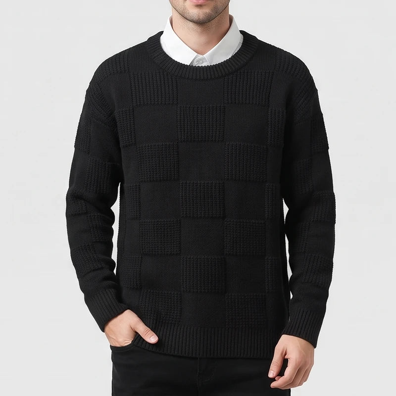 Man wearing a black knit sweater with a textured checkerboard pattern over a white collared shirt, hand in pocket.